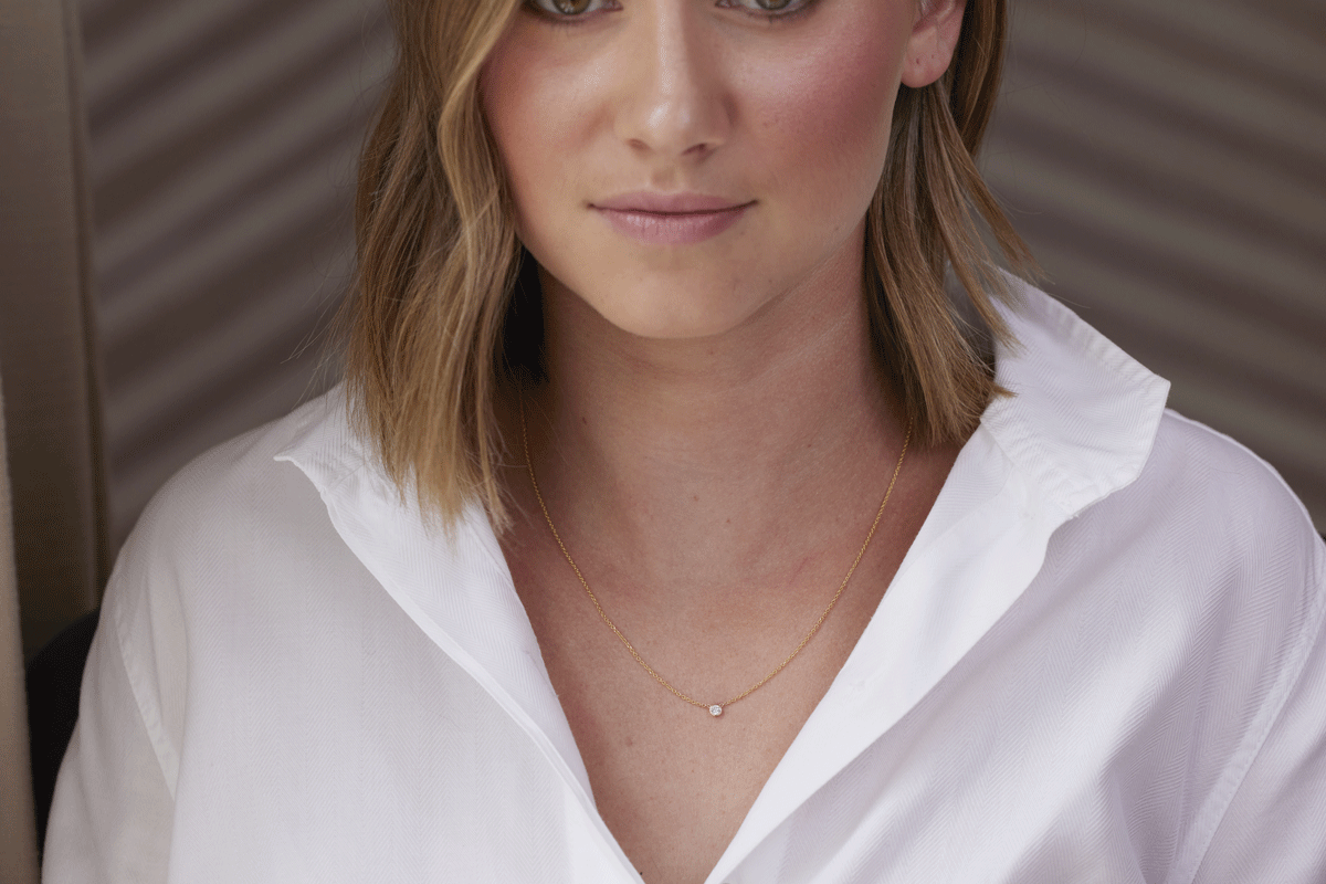 Young female with a white collared shirt and thin gold chain necklace with singular round diamond baguette
